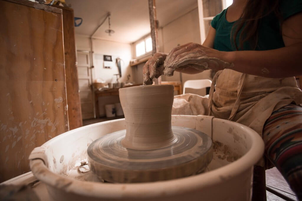 Una persona con delantal da forma a una vasija de barro en un torno de alfarero en un taller. La habitación, ligeramente abarrotada de equipos de cerámica, refleja el espíritu artístico de este espacio de inicio creativo.