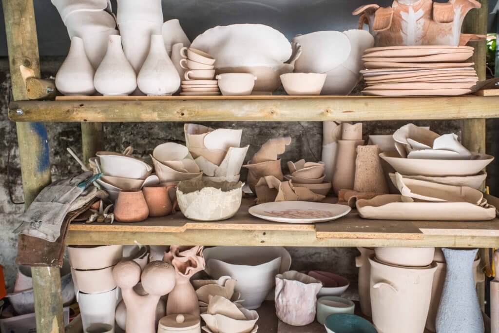 Un estante de madera contiene varias piezas de cerámica cruda y cocida, incluidos cuencos, jarrones, platos y jarras de diversas formas y tamaños de clases de cerámica recientes.