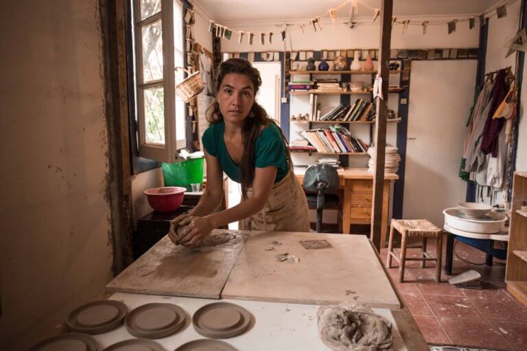 Una persona amasa arcilla sobre una mesa en un taller de cerámica, con varias herramientas, libros y piezas de cerámica de fondo. Este espacio creativo encarna el espíritu de colaboración y expresión artística que define a Nosotros.