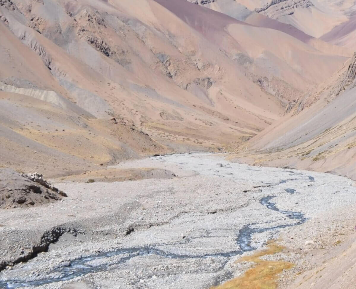 Un valle seco y rocoso con escasa vegetación y un pequeño arroyo sinuoso que recuerda a los paisajes de Grof. Montañas y crestas áridas rodean la zona bajo un cielo despejado.