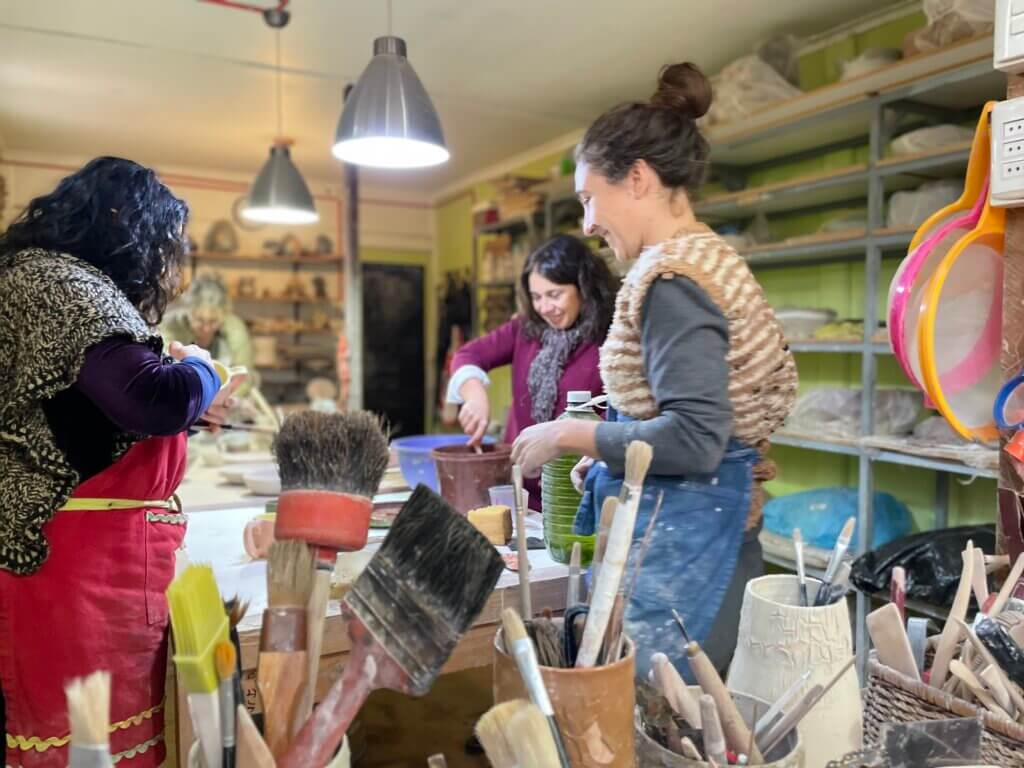Tres personas trabajan en proyectos de cerámica en un estudio bien iluminado y lleno de estanterías con materiales y herramientas, lo que crea un ambiente perfecto para el aprendizaje. En primer plano se ven pinceles que enfatizan el aspecto educativo de sus clases.
